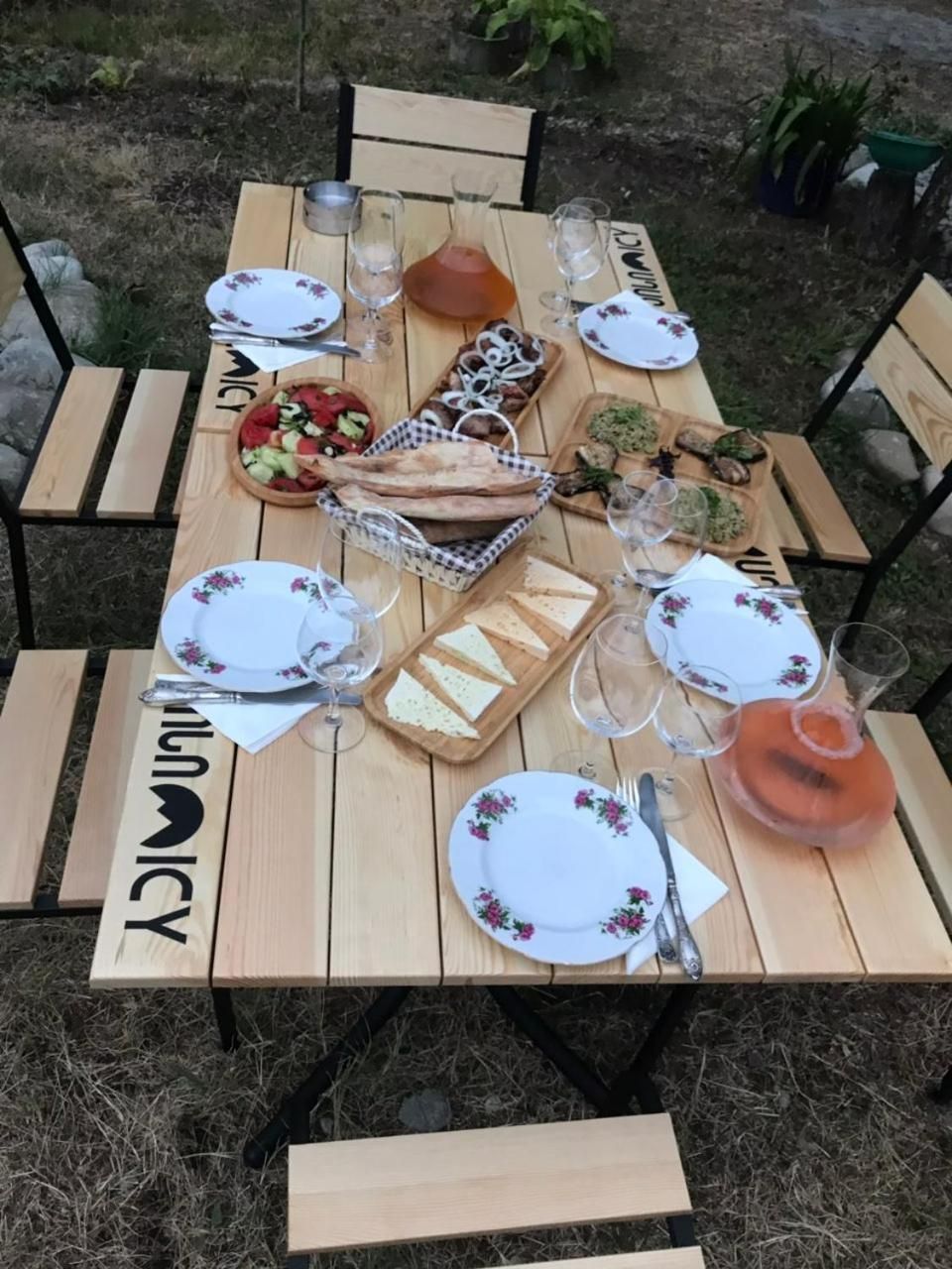 An outdoor wooden table set for a meal with plates, cutlery, glasses, and a variety of food items.