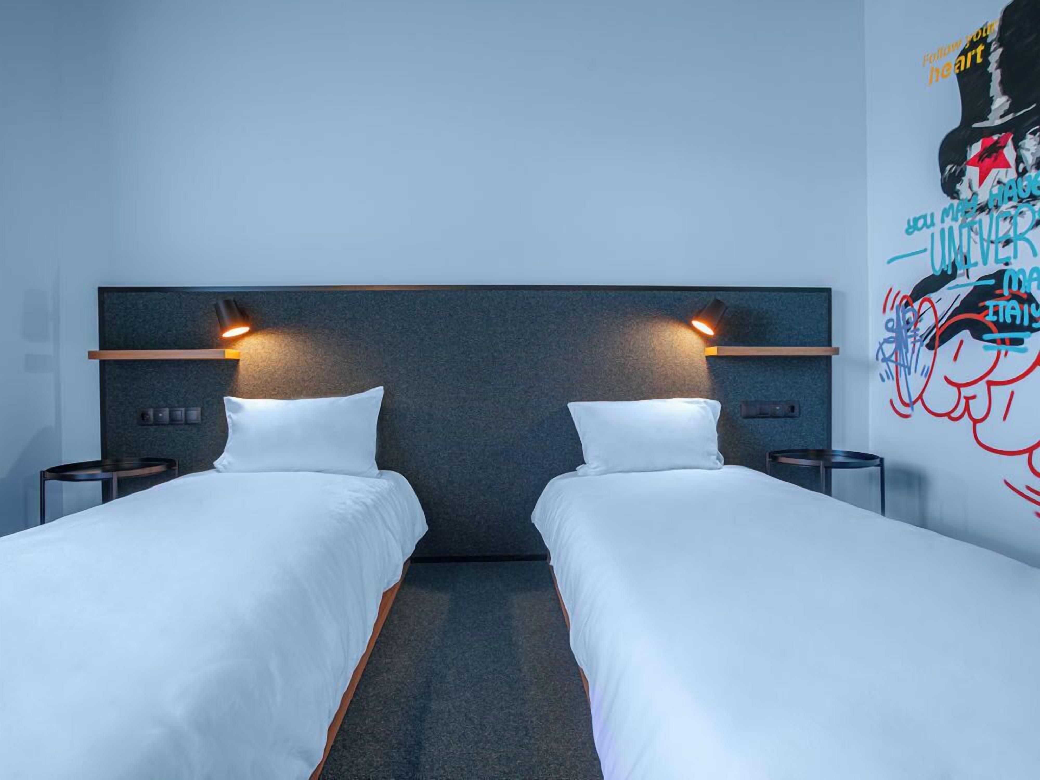 Ein Hotelzimmer mit zwei Einzelbetten, einem dunklen Kopfteil, Nachttischlampen, einem kleinen Tisch und einer Wand mit einem farbenfrohen Wandgemälde.