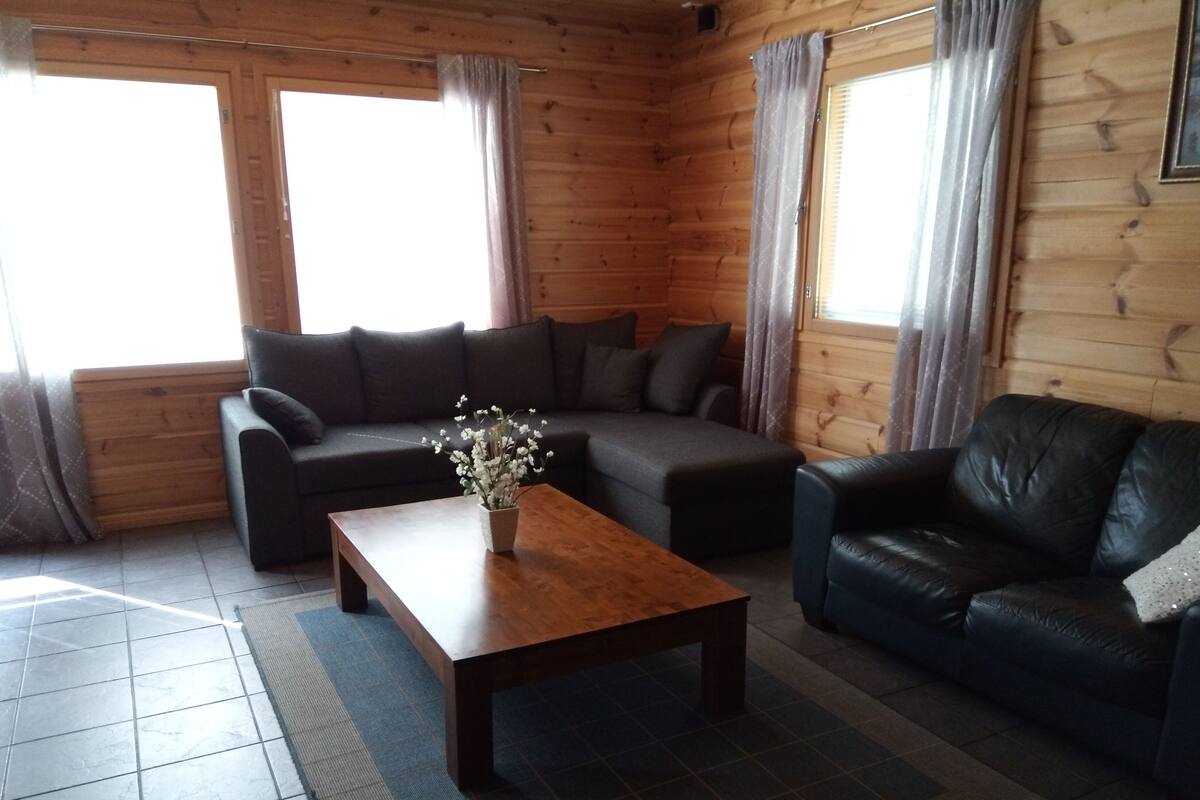 A wooden cabin interior with a dark brown sofa, a black leather couch, and a wooden coffee table with a white flower arrangement.