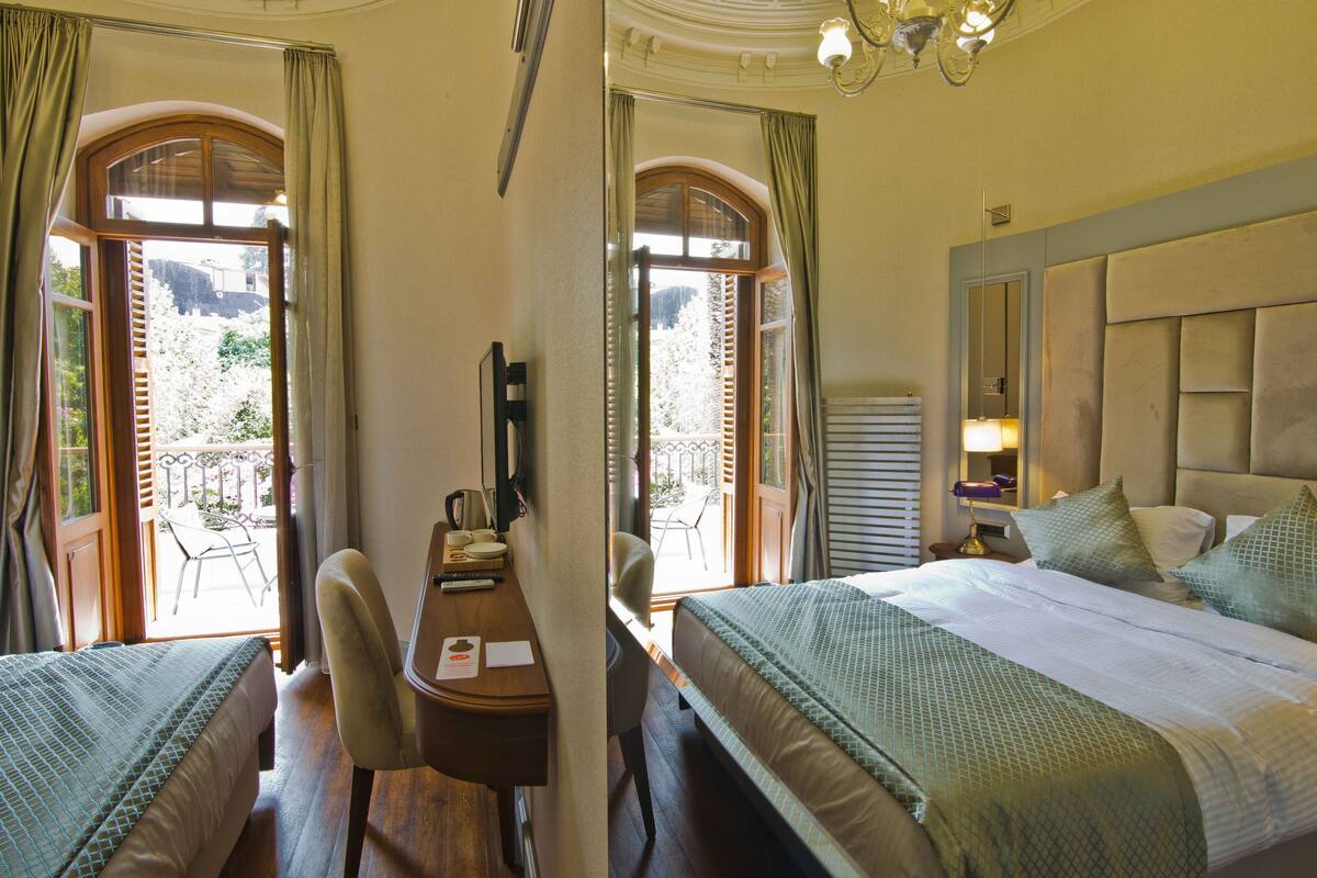 Habitación de hotel con dos camas, un escritorio y una silla. Se ve un balcón con mesa y sillas a través de las ventanas.