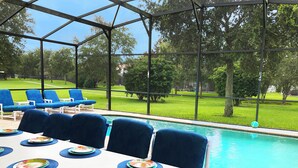 Ein Essbereich am Pool mit einem Tisch für vier Personen, blauen Sitzgelegenheiten und Blick auf Pool und Rasen.