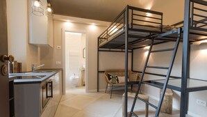 Una cocina compacta con lavamanos integrado y una pequeña zona de comedor. Se observa una cama litera con escalera y una silla de mimbre al fondo.