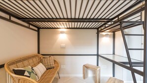 Una habitación con techo alto, un sofá de ratán con cojines, un pequeño banco de madera y una escalera metálica.