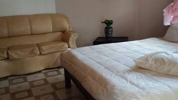 A bedroom with a beige sofa, a bed with white bedding, a small table with a vase, and a hanging pink fabric.