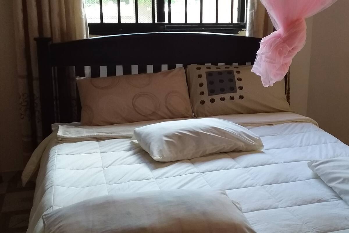 Un lit bien fait, avec une couette et des oreillers blancs, une tête de lit sombre et une fenêtre équipée de stores.