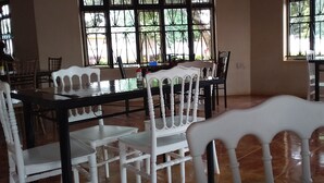 Un área de comedor con sillas blancas y una mesa negra, ventanas amplias con barrotes de metal y piso de baldosas.