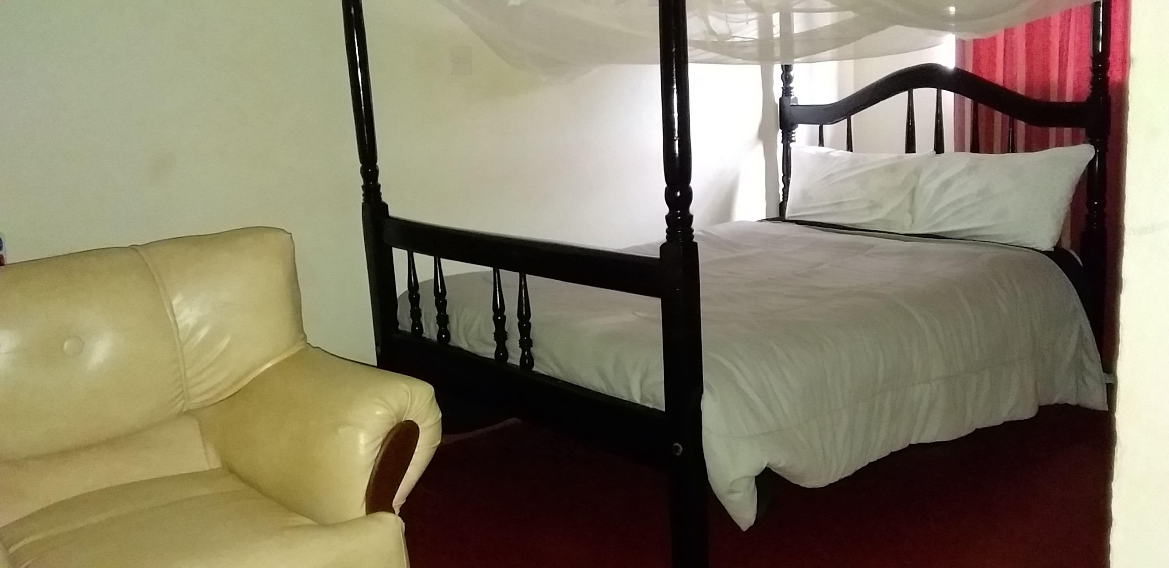 Een kamer met een hemelbed met witte lakens, een leren fauteuil en een rood gordijn, met witte wanden.