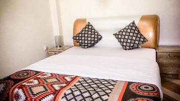 Una cama con una colcha estampada, dos almohadas con diseño geométrico y una mesita de noche con una lámpara.