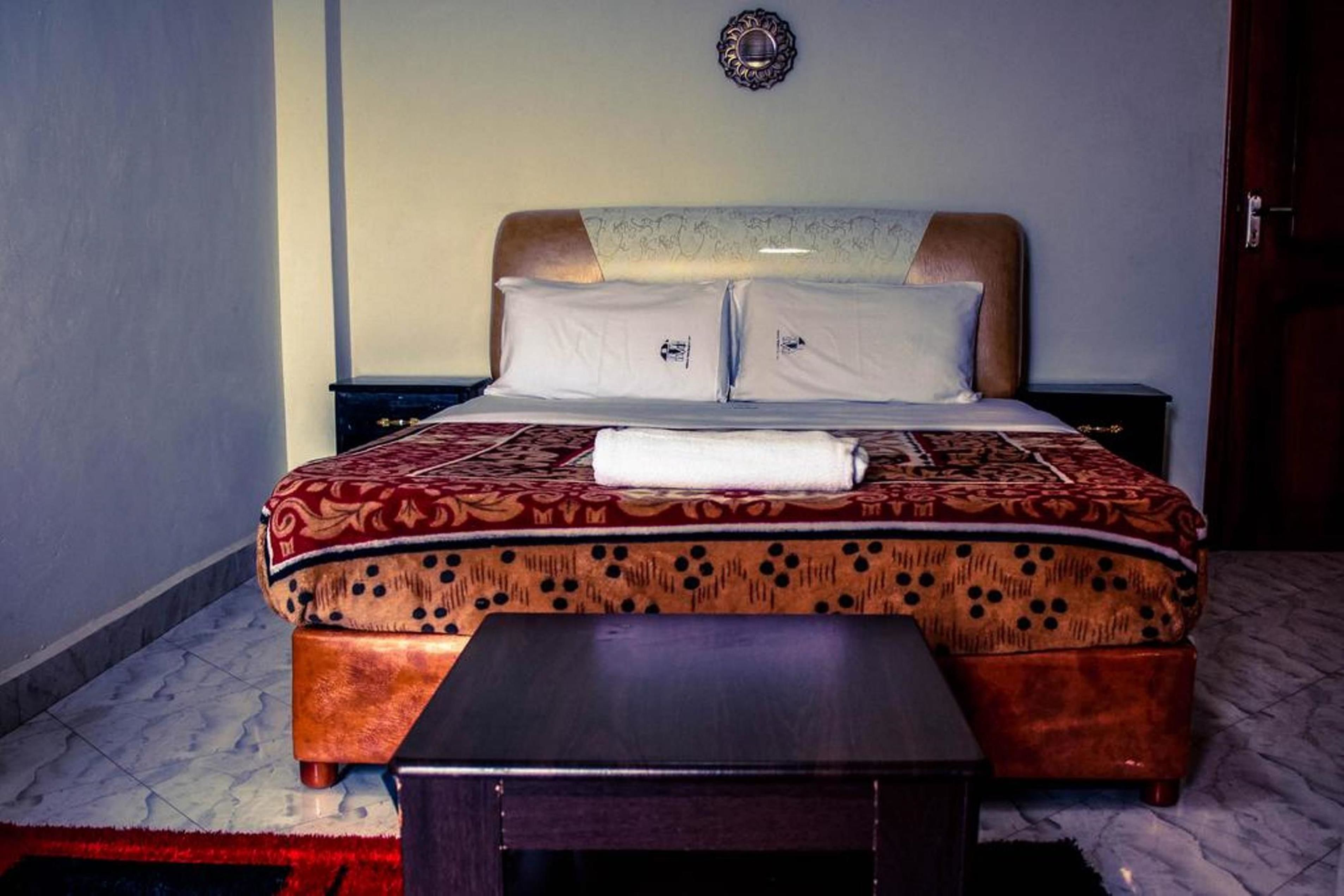 A neatly made bed with a patterned bedspread and pillows, a wooden nightstand, and a small table in a hotel room.