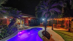Una piscina de borde curvo, rodeada de palmeras y un patio con sillas y mesas.