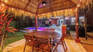 Un área de comedor al aire libre con techo de paja, mesas y sillas de madera.