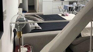 Quarto moderno e compacto, com cama, mesa de cabeceira e pequena escrivaninha com cadeira. Uma escada leva a uma área de cozinha com armários e pia.