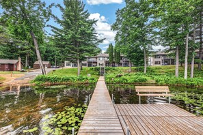A wooden dock leading to a pond with lily pads, surrounded by trees and a building with multiple balconies. - Spacious Black Bear lakefront condo with private dock! (St Germain)