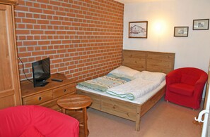 Ein Zimmer mit Backsteinwand, einem hölzernen Bett, einem roten Stuhl, einem Holztisch mit Fernseher und einem kleinen Hocker.
