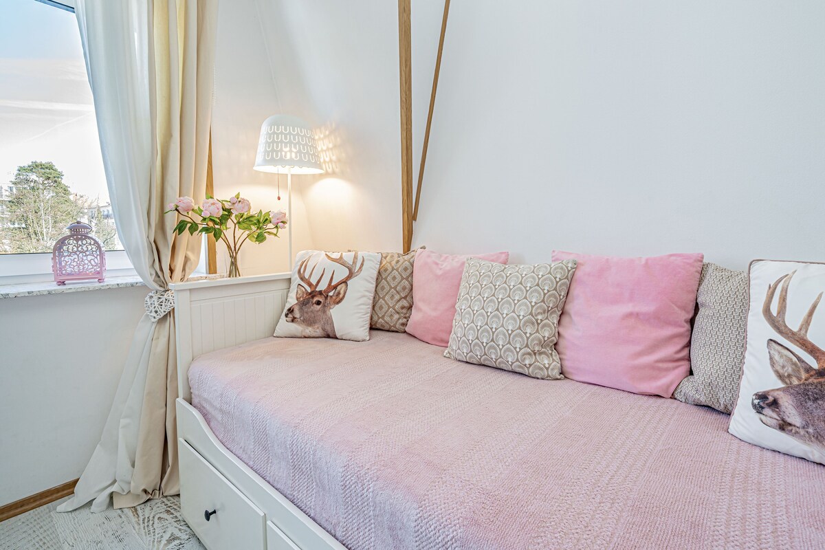 Quarto com cama, almofadas, mesa de cabeceira, abajur e janela com cortinas.