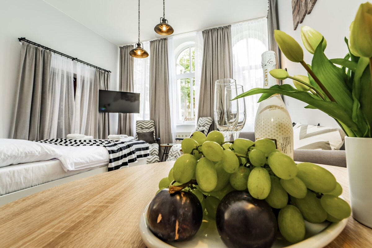 Quarto de hotel com uma cama, uma TV, uma mesa de jantar com uvas e uma garrafa, e uma planta em vaso.