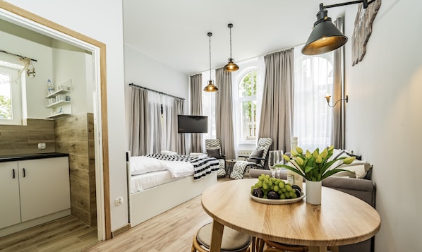 A modern hotel room with a kitchenette, a bed with a checkered duvet, a round dining table with a plant and fruit, and a TV mounted on the wall.