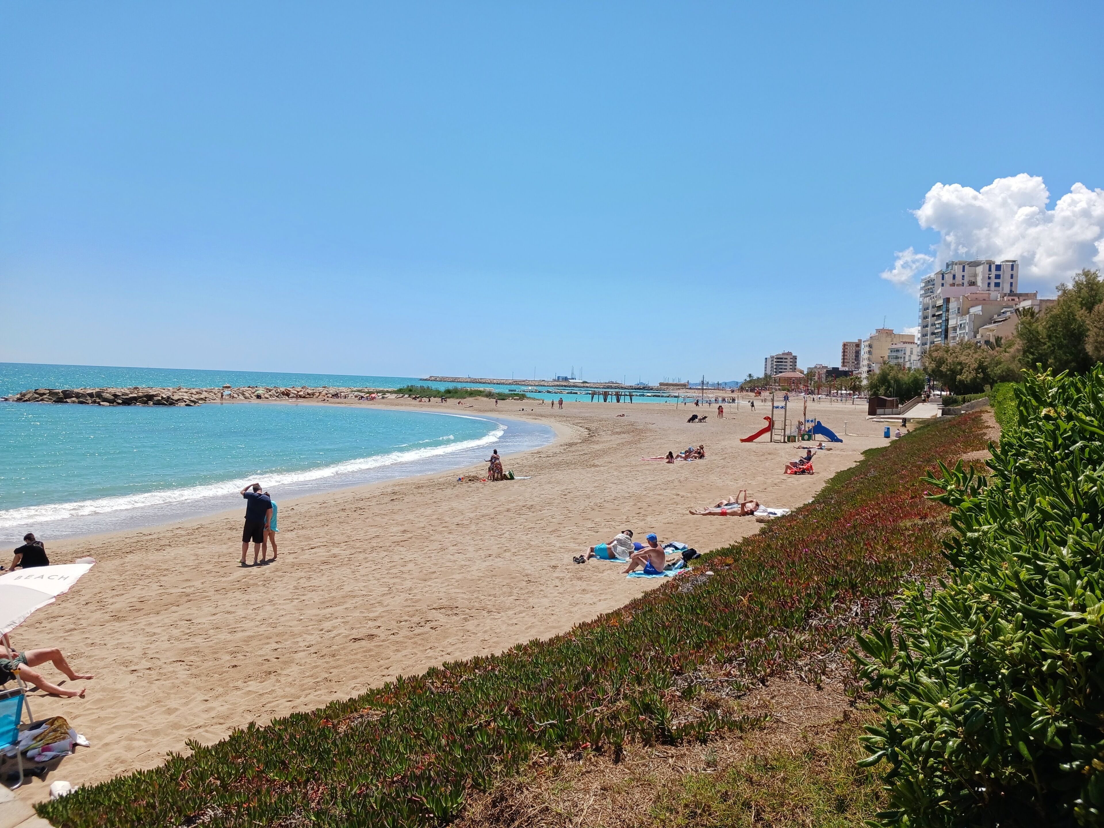 Une plage où l’on voit des gens se prélasser au soleil et nager, une digue rocheuse et des bâtiments en arrière-plan.