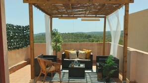 Un espacio de estar al aire libre techado con una pérgola de madera, un sofá con cojines amarillos, una silla de mimbre y una mesita con una botella y un vaso.