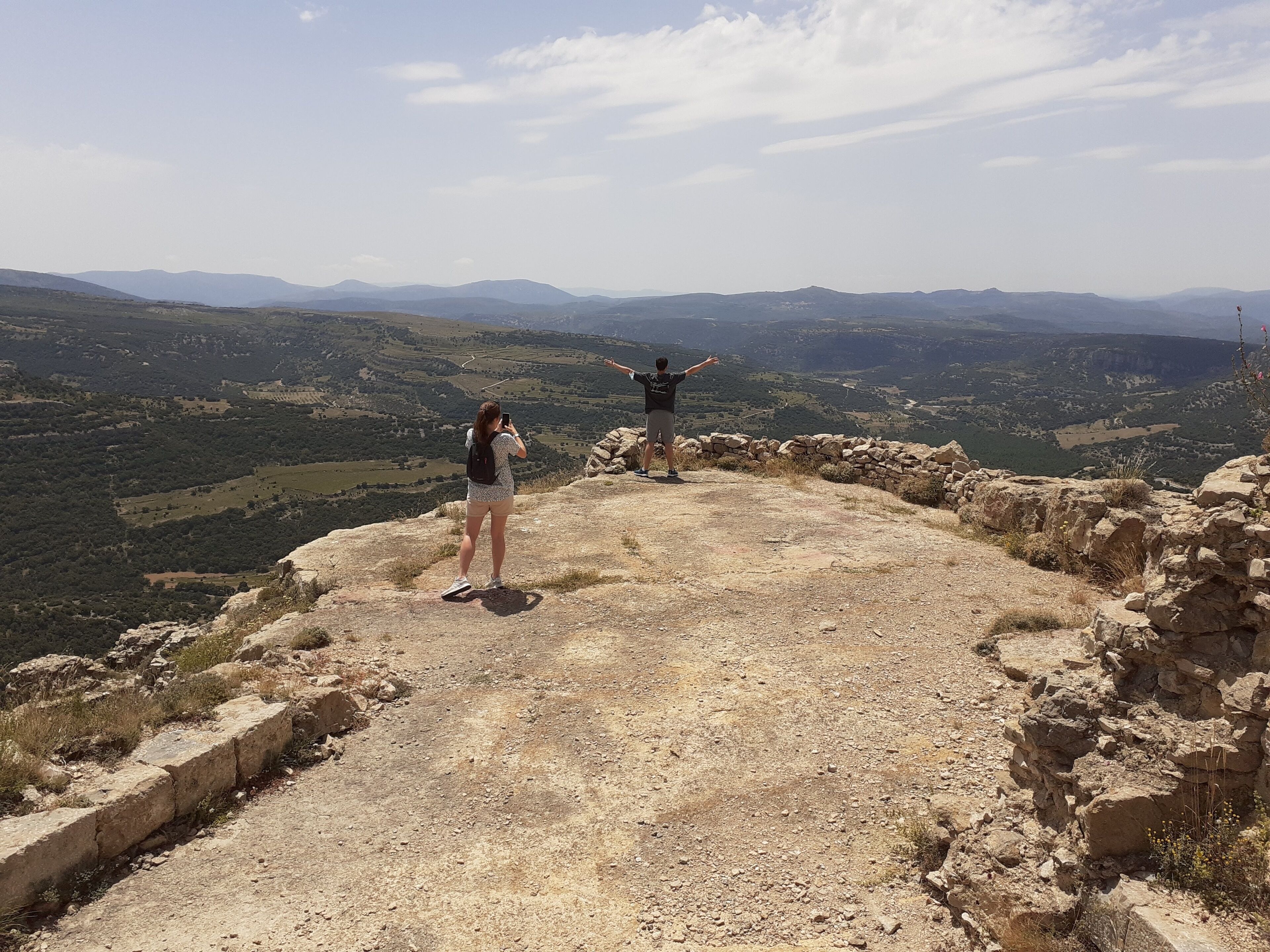 Un terrain rocheux, avec un mur de pierres, où se tient une personne, les bras tendus, surplombant une vallée et des montagnes lointaines.