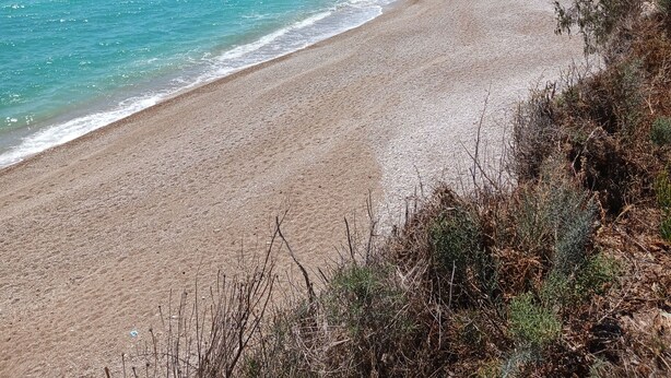Una playa con agua turquesa cristalina, una orilla arenosa y algunos edificios al fondo.