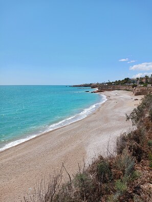 A beach with clear turquoise water, a sandy shore, and a few buildings in the background.