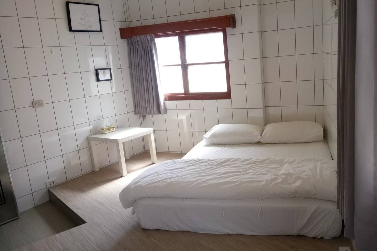 Un dormitorio con una cama, una mesita de noche, una ventana con cortinas y un muro con baldosas.