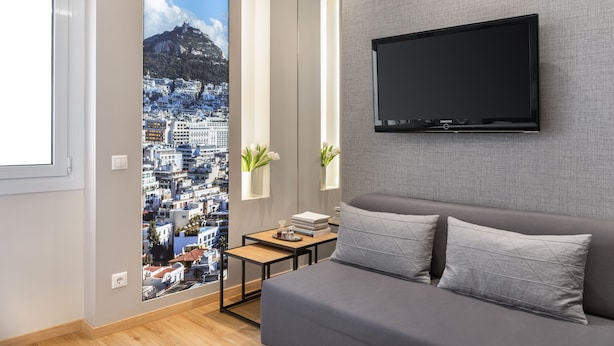 Moderni hotellihuone, jossa on suuri ikkuna, seinälle asennettu taulutelevisio, harmaa sohva sekä pieni pöytä, jonka päällä on kirjoja ja maljakko kukkia.