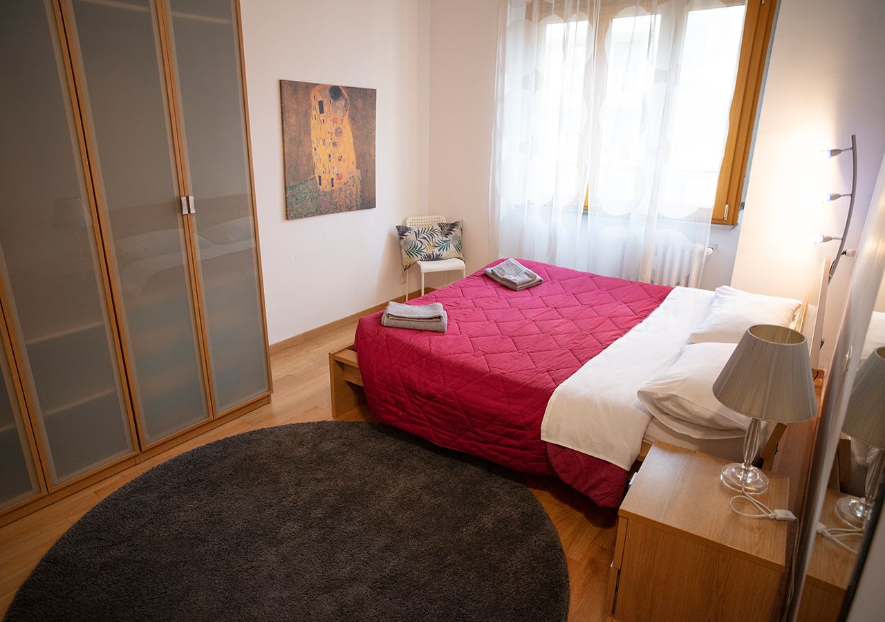 A bedroom with a bed, a bedside table with a lamp, a chair, a painting on the wall, and a large rug.