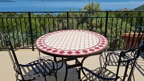 A round table with a patterned tablecloth, surrounded by metal chairs, set on a balcony with a view of a large body of water and distant hills. - Appart'hotel Résidence La Provence (Saint-Mitre-les-Remparts)