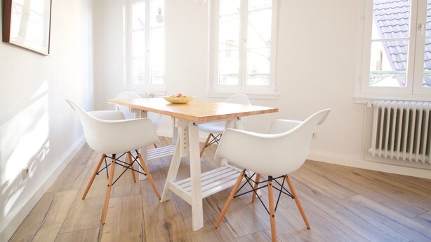 Un comedor luminoso con una mesa de madera y sillas blancas, ventanas amplias y un radiador.