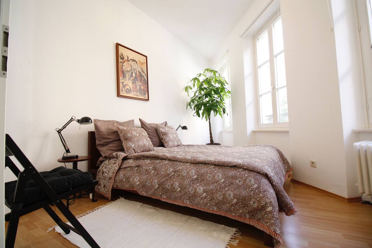 A bedroom with a bed, a desk, a chair, a lamp, a plant, and a window.
