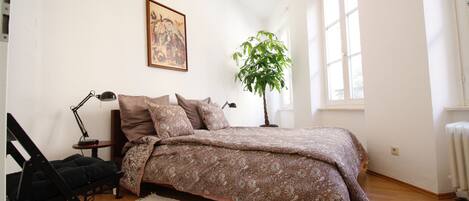 Ein Schlafzimmer mit Bett, Schreibtisch, Stuhl, Lampe, Pflanze und Fenster.