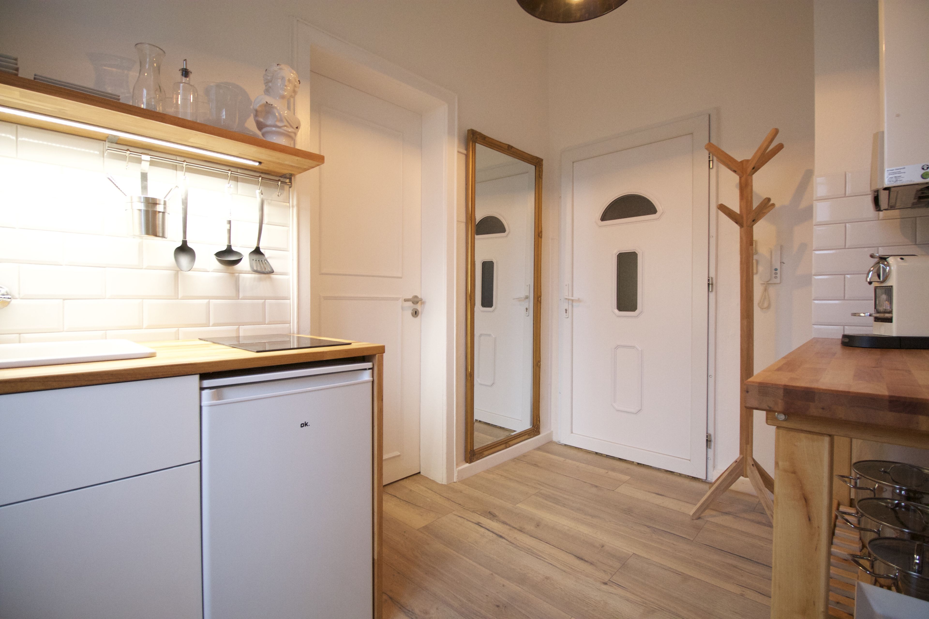 A modern kitchen with white cabinetry, a wooden countertop, and a built-in dishwasher. There is a coat rack and a mirror on the wall.
