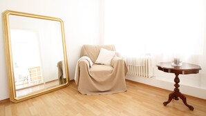 Una habitación con un espejo grande, un sillón color beige, una mesita redonda y un radiador.