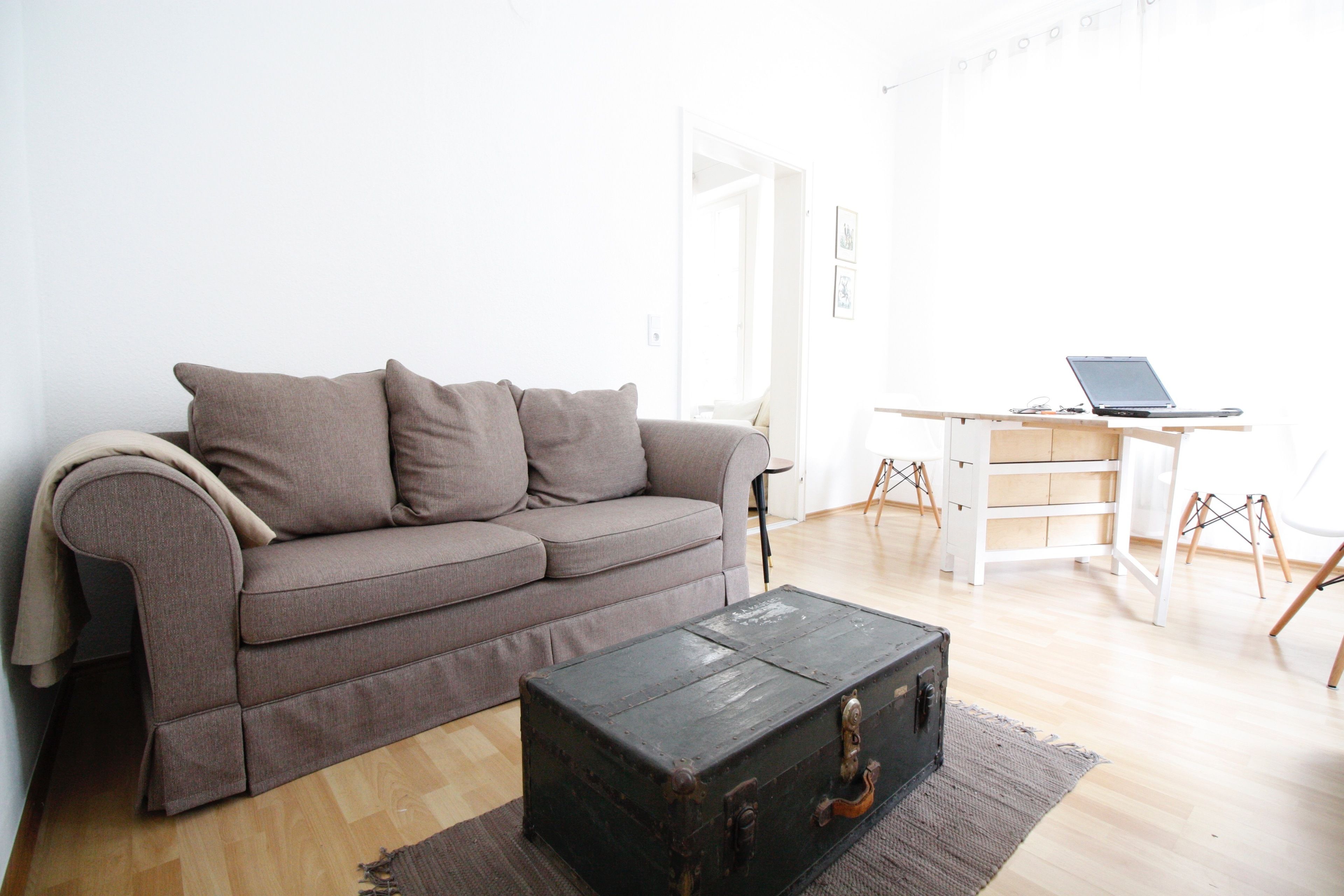 A living room with a sofa, a wooden floor, a small table, and a laptop on a desk.