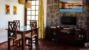 Un comedor con una mesa de vidrio y sillas de madera, una pared de piedra con un televisor, y un cuadro sobre el televisor.
