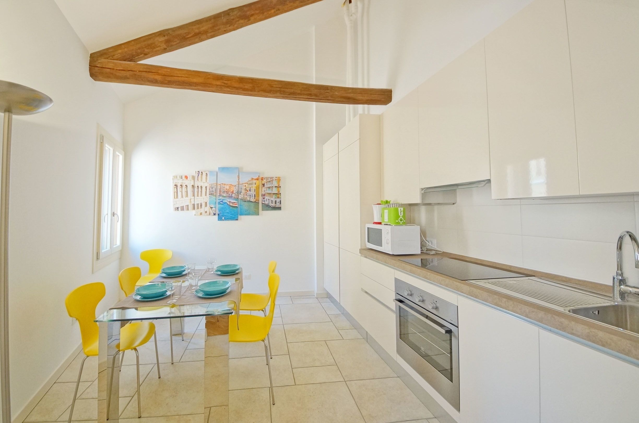 A compact kitchen with white cabinets, a microwave, and a sink. There is a dining area with yellow chairs and a glass table.