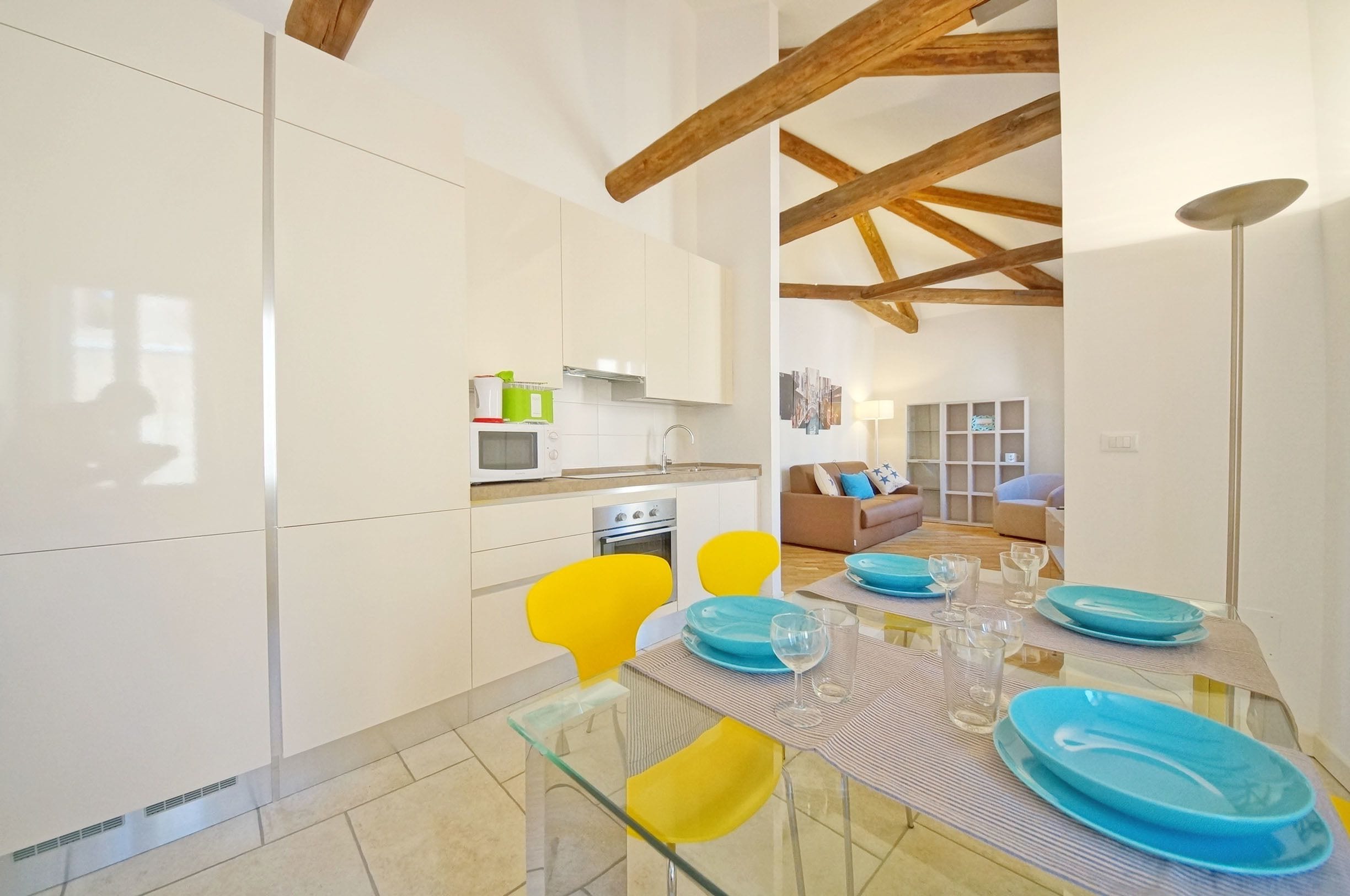 Une cuisine moderne avec des meubles blancs, un four à micro-ondes et une cuisinière. On y trouve un coin repas avec une table en verre, des chaises jaunes et des assiettes bleues.
