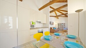 A modern kitchen with white cabinets, a microwave, and a stove. There is a dining area with a glass table, yellow chairs, and blue plates. - DolceVita Apartments N. 394 (Venice)
