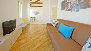 A modern living room with a flat-screen TV, wooden flooring, a brown sofa with blue pillows, and a dining area in the background. - DolceVita Apartments N. 394 (Venice)