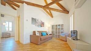 A spacious living room with wooden flooring, a brown sofa with blue cushions, a white armchair, a TV on a cabinet, and a wall with framed pictures. - DolceVita Apartments N. 394 (Venice)