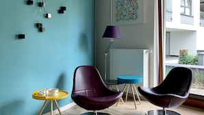 Un salon moderne avec des murs de couleur turquoise, une grande fenêtre, deux fauteuils violets, une table jaune et un lampadaire.