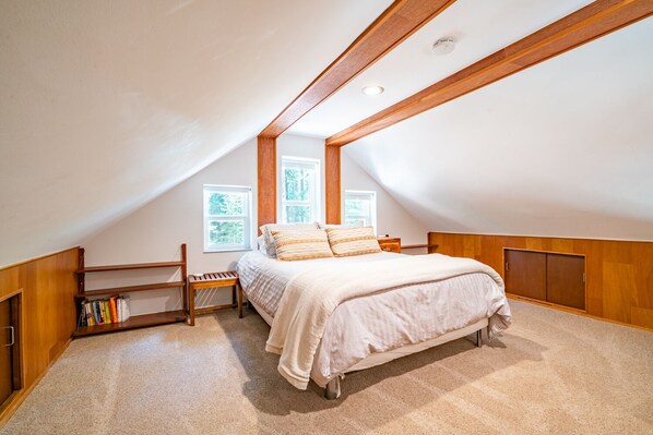 Un dormitorio con techo inclinado, una cama con colcha color beige, un banco de madera y una estantería.