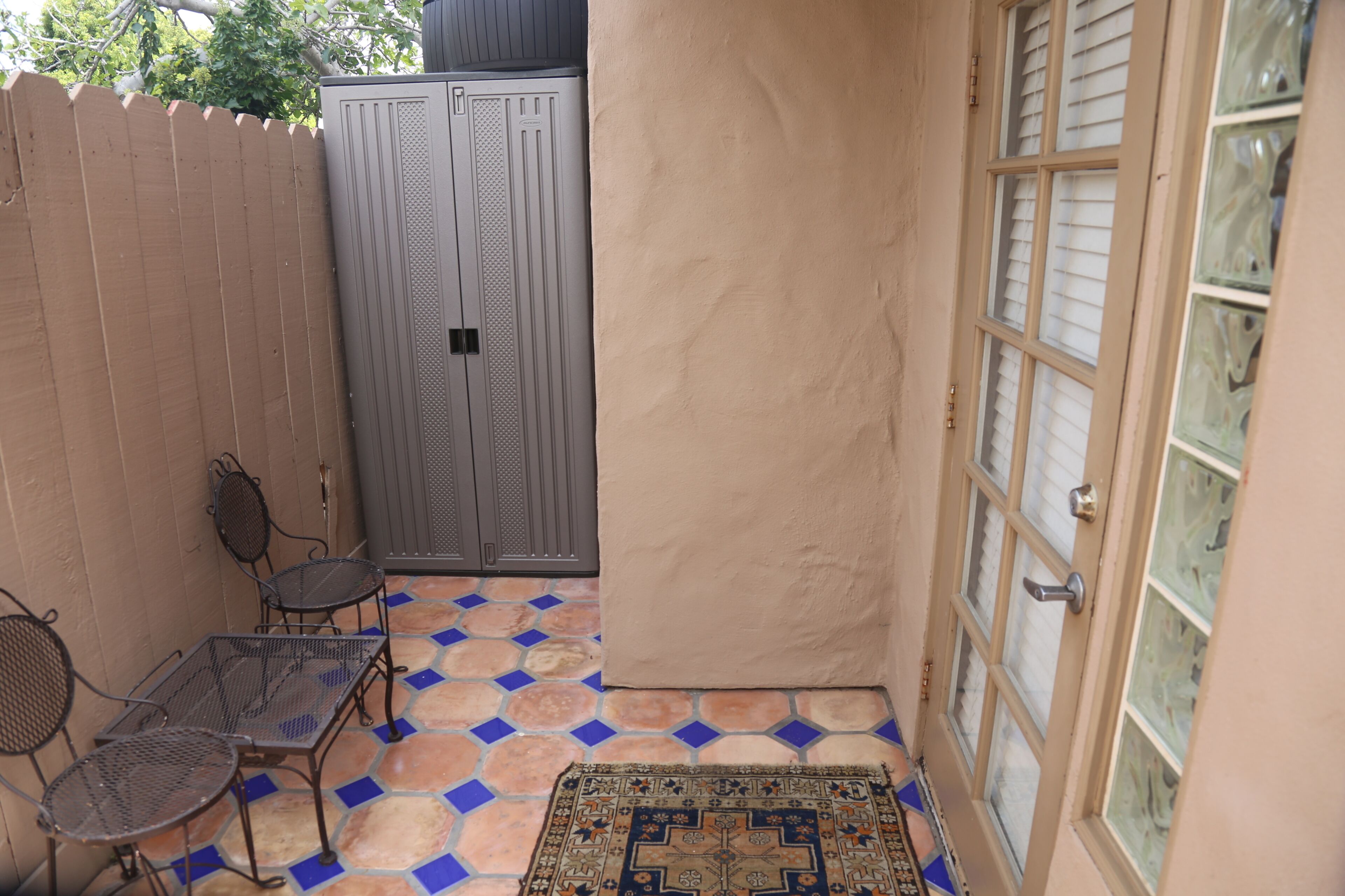 Un pequeño patio con una mesa y sillas de metal, un tapiz estampado y una puerta con ventanal.