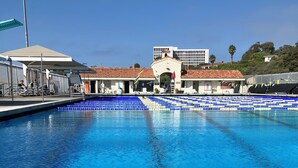 En swimmingpool med blåt vand, et overdækket siddeområde og en bygning med et tagtækket tag i baggrunden.