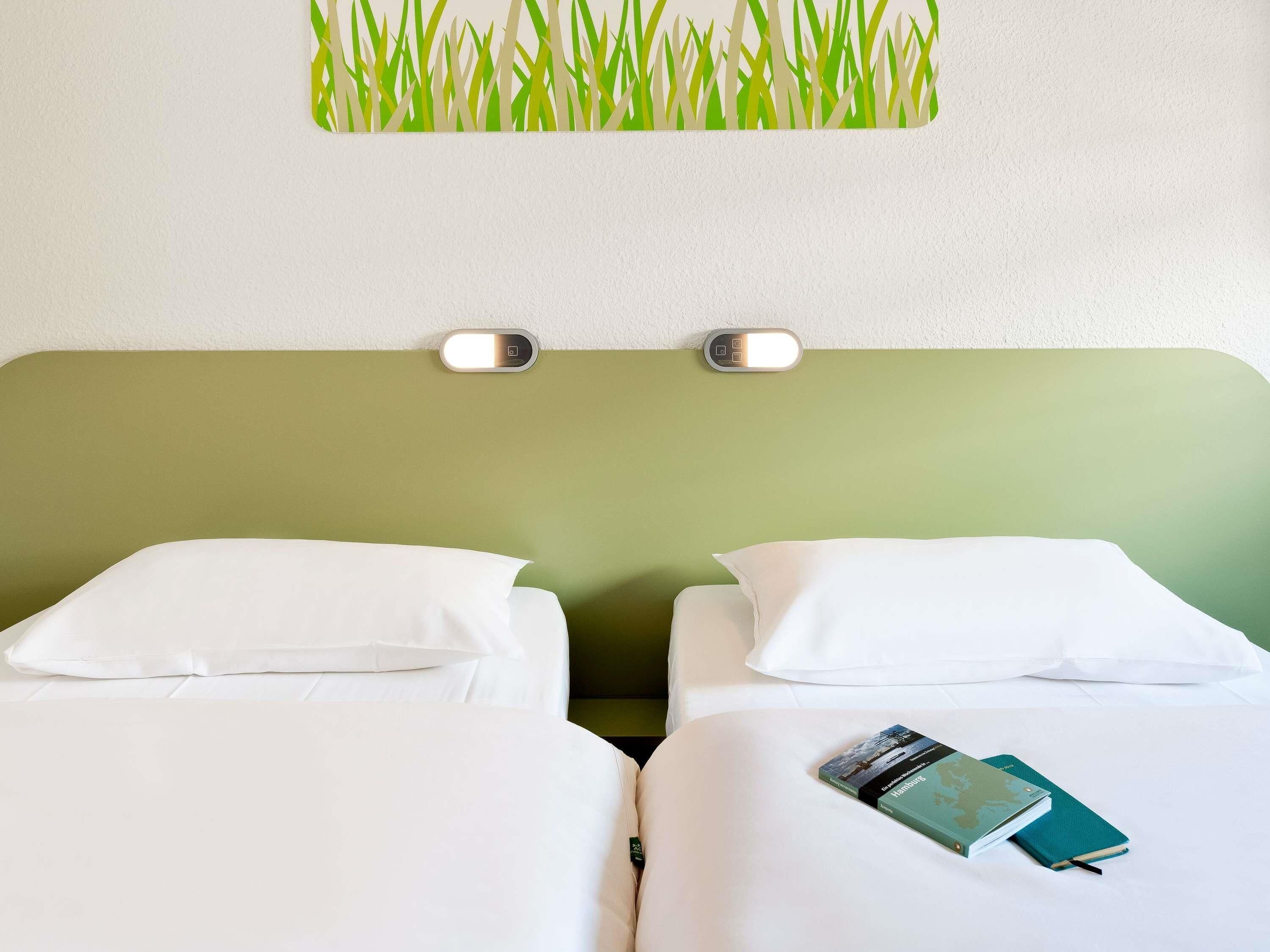 A hotel room with two beds, white bedding, a green headboard, and a wall-mounted light fixture.