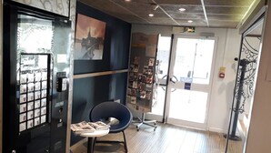 Una habitación con puerta de vidrio, una silla azul, una mesita con revistas y una estantería con diversos objetos.