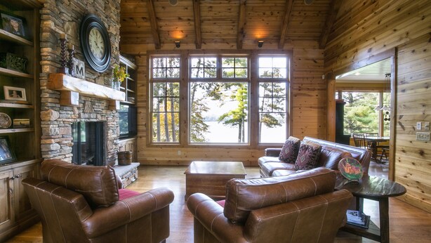 Interior acogedor de una cabaña de troncos con sofás de cuero, una chimenea de piedra y amplias ventanas con vistas a los árboles.