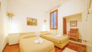 A hotel room with two beds, a wooden floor, a window with curtains, a chair with a patterned cushion, and a painting on the wall. - DolceVita Apartments N. 408 (Venice)